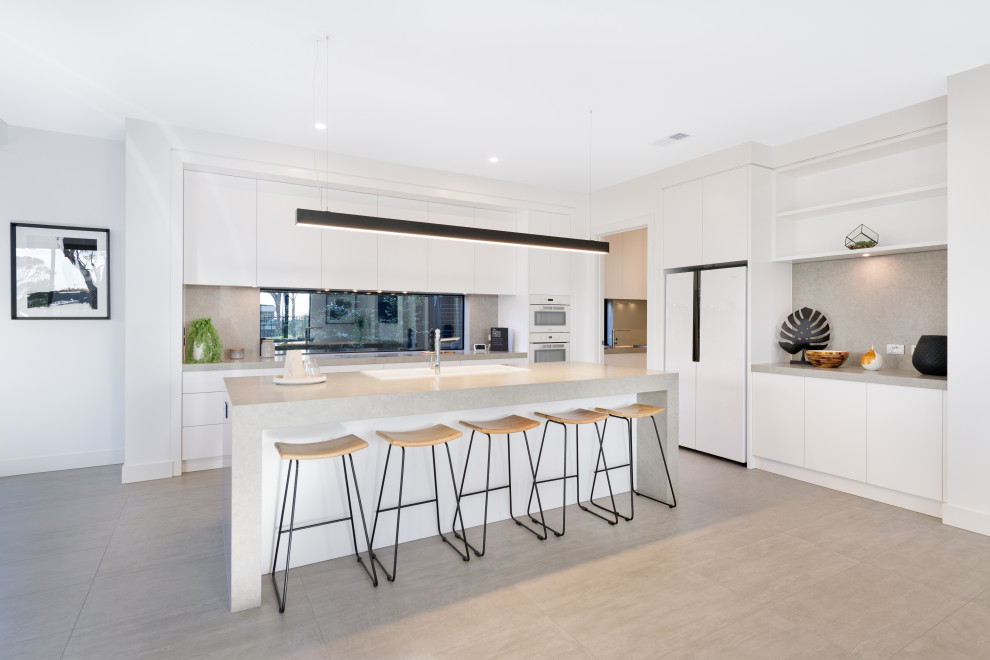 This is an example of a large contemporary l-shaped kitchen in Melbourne with a drop-in sink, flat-panel cabinets, white cabinets, grey splashback, white appliances, with island, grey floor and grey benchtop.