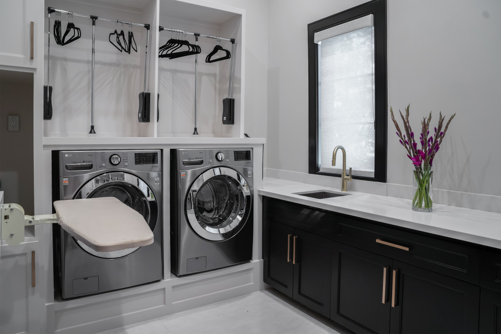 Photo of a large transitional u-shaped dedicated laundry room with an undermount sink, shaker cabinets, black cabinets, solid surface benchtops, white walls, porcelain floors, an integrated washer and dryer, white floor and white benchtop.