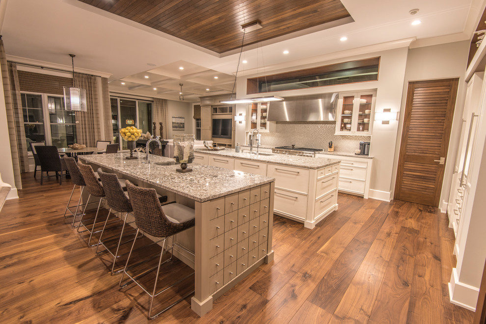 Photo of a large tropical u-shaped open plan kitchen in Tampa with an undermount sink, shaker cabinets, white cabinets, granite benchtops, grey splashback, glass tile splashback, panelled appliances, light hardwood floors and multiple islands.