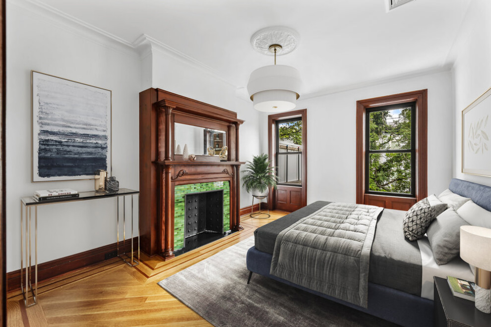 Expansive traditional guest bedroom in New York with white walls, light hardwood flooring, a standard fireplace, a wooden fireplace surround and beige floors.