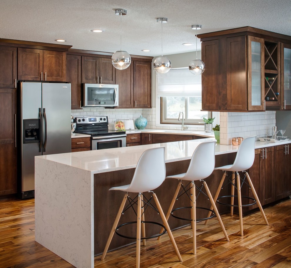 This is an example of a mid-sized transitional l-shaped kitchen in Omaha with shaker cabinets, brown cabinets, quartz benchtops, white splashback, subway tile splashback, stainless steel appliances, medium hardwood floors, a peninsula and brown floor.