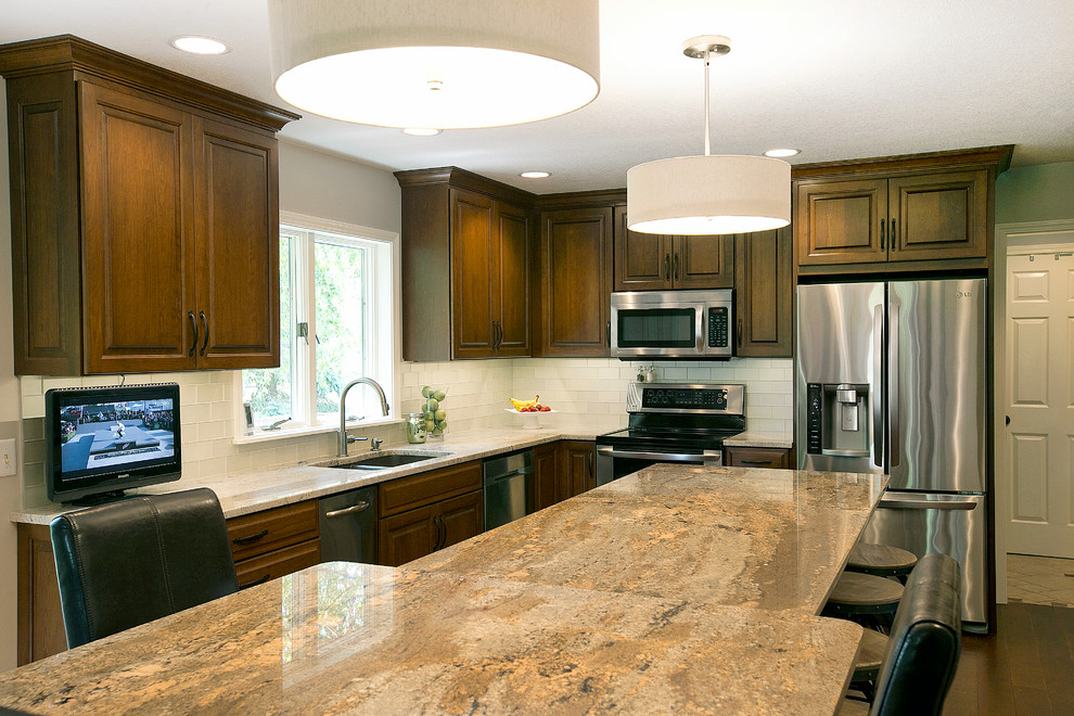 Transitional l-shaped medium tone wood floor eat-in kitchen photo with a double-bowl sink, raised-panel cabinets, dark wood cabinets, granite countertops, white backsplash, glass tile backsplash, stainless steel appliances and an island