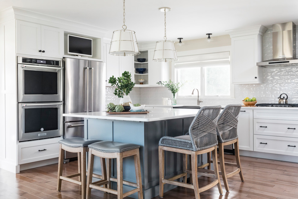 Large beach style l-shaped eat-in kitchen in Boston with a farmhouse sink, shaker cabinets, white cabinets, quartz benchtops, white splashback, glass tile splashback, stainless steel appliances, with island, white benchtop, light hardwood floors and brown floor.