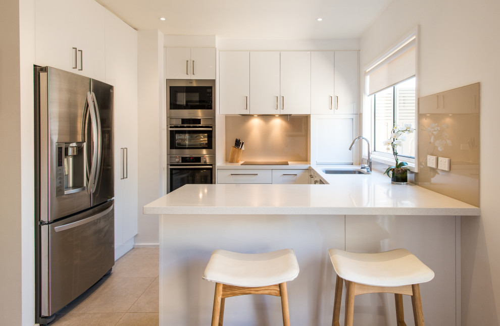 Photo of a contemporary u-shaped kitchen in Melbourne with an undermount sink, flat-panel cabinets, white cabinets, beige splashback, stainless steel appliances, a peninsula, brown floor and white benchtop.