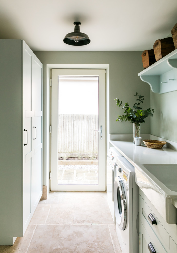 This is an example of a beach style single-wall dedicated laundry room in Sussex with a farmhouse sink, shaker cabinets, green cabinets, green walls, a side-by-side washer and dryer, beige floor and white benchtop.