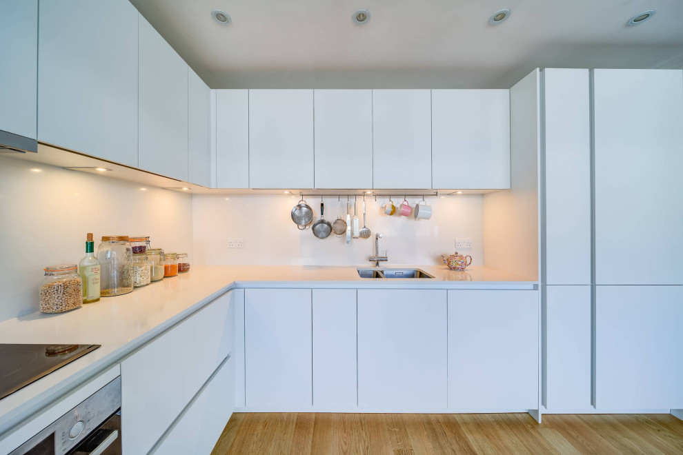 Inspiration för ett mellanstort funkis vit vitt kök, med en dubbel diskho, släta luckor, vita skåp, bänkskiva i kvartsit, vitt stänkskydd, svarta vitvaror, ljust trägolv och brunt golv