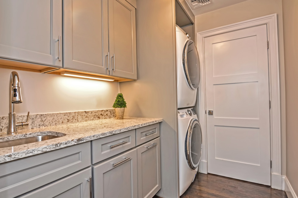 This is an example of a small transitional galley dedicated laundry room in Boston with an undermount sink, shaker cabinets, grey cabinets, quartz benchtops, grey splashback, engineered quartz splashback, beige walls, dark hardwood floors, a stacked washer and dryer, grey floor and grey benchtop.