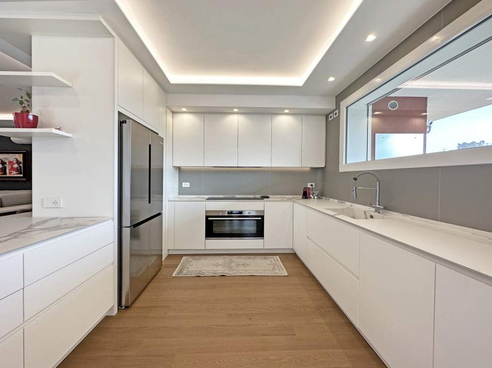Example of a large minimalist u-shaped medium tone wood floor, brown floor and tray ceiling eat-in kitchen design in Milan with an integrated sink, flat-panel cabinets, white cabinets, marble countertops, white backsplash, cement tile backsplash, stainless steel appliances, an island and white countertops