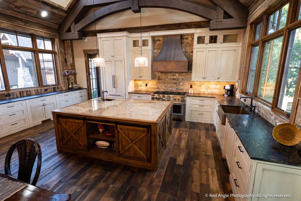 Example of a large mountain style dark wood floor and exposed beam kitchen design with shaker cabinets, distressed cabinets, granite countertops, stone tile backsplash, stainless steel appliances and an island