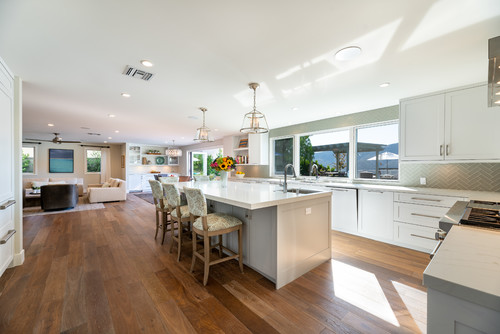 Open-concept modern kitchen with white kitchen cabinets, large island with bar stools, wood flooring, and panoramic windows connecting to a bright living area.