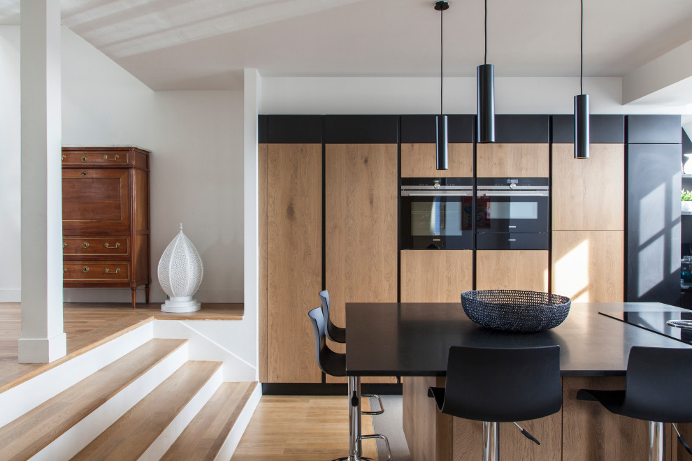 Idées déco pour une grande cuisine ouverte contemporaine en L et bois clair avec un placard à porte plane, un sol en bois brun, îlot, plan de travail noir, un évier encastré, une crédence noire, une crédence en marbre, un électroménager noir et un sol gris.