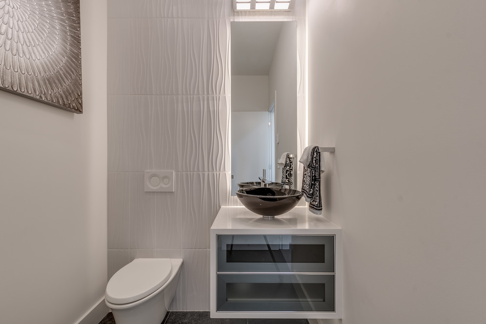 Small modern powder room in Vancouver with glass-front cabinets, grey cabinets, a wall-mount toilet, white tile, porcelain tile, white walls, ceramic floors, a vessel sink, engineered quartz benchtops, grey floor and white benchtops.