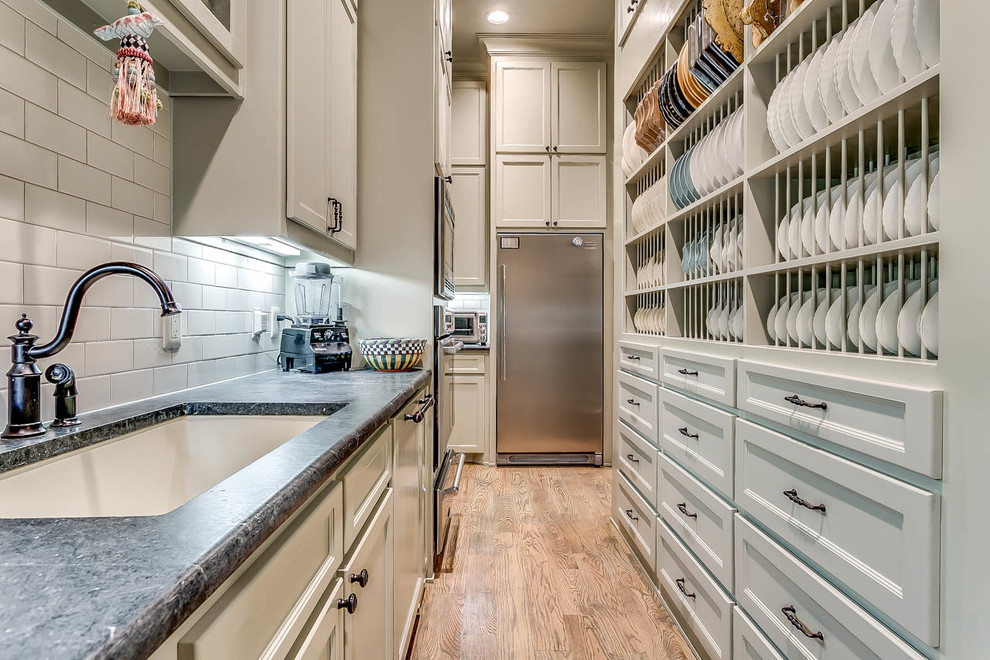 Design ideas for a large traditional galley kitchen pantry in Dallas with medium hardwood floors, an undermount sink, recessed-panel cabinets, grey cabinets, soapstone benchtops, white splashback, ceramic splashback, stainless steel appliances and no island.