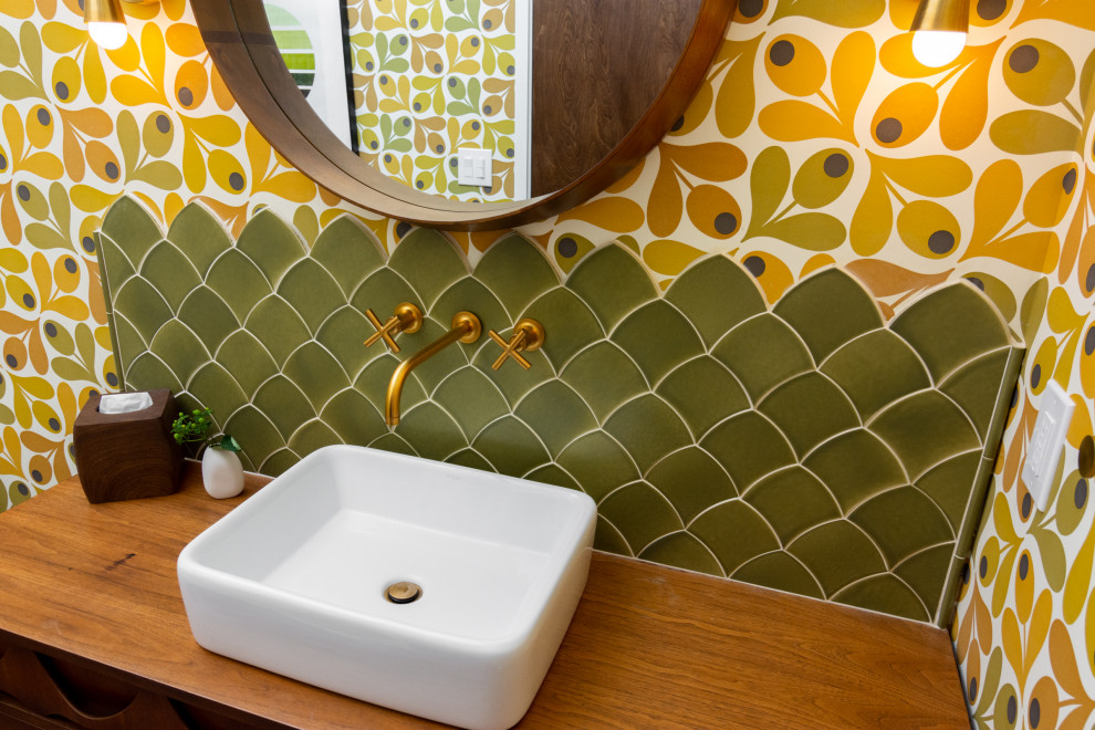 This is an example of a mid-sized midcentury powder room in Detroit with furniture-like cabinets, brown cabinets, a one-piece toilet, green tile, ceramic tile, multi-coloured walls, light hardwood floors, a vessel sink, wood benchtops, brown floor, brown benchtops, a freestanding vanity, vaulted and wallpaper.