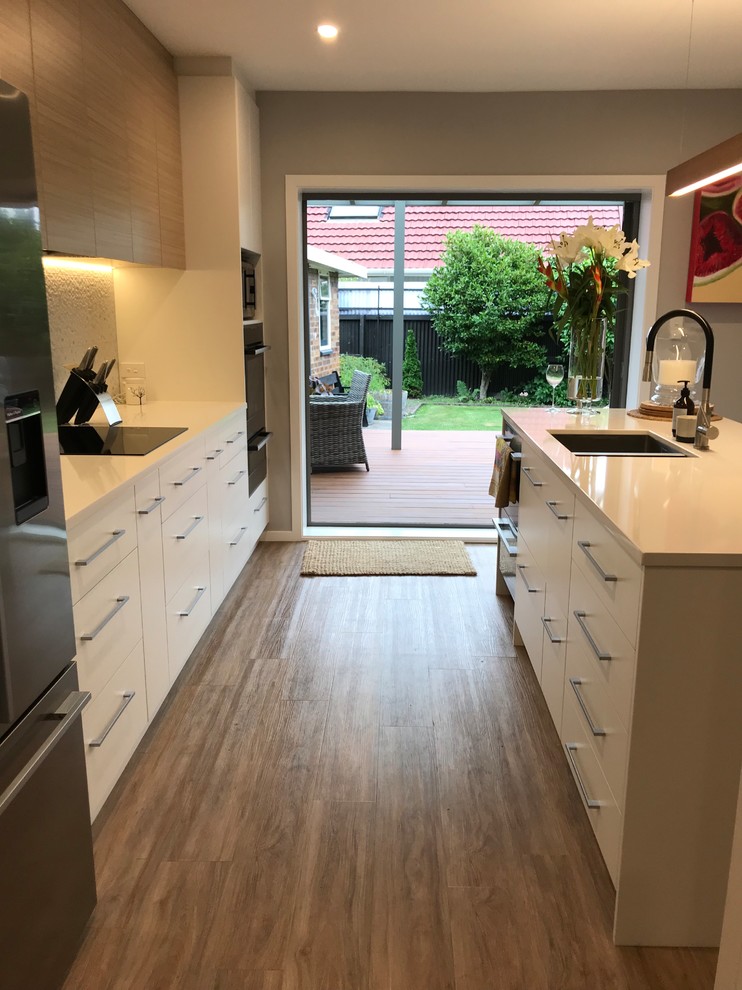 Inspiration for a large scandinavian galley separate kitchen in Christchurch with a double-bowl sink, flat-panel cabinets, white cabinets, quartz benchtops, grey splashback, porcelain splashback, stainless steel appliances, vinyl floors, with island and brown floor.