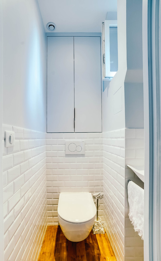 This is an example of a small transitional powder room in Paris with beaded inset cabinets, blue cabinets, a wall-mount toilet, white tile, subway tile, blue walls, light hardwood floors, a vessel sink and brown floor.