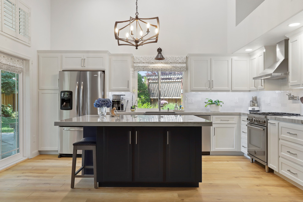 This is an example of a contemporary eat-in kitchen in San Francisco with a farmhouse sink, shaker cabinets, white cabinets, quartzite benchtops, white splashback, ceramic splashback, stainless steel appliances, with island, brown floor and white benchtop.