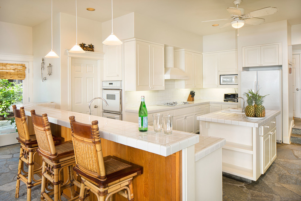 Large beach style u-shaped eat-in kitchen in Hawaii with recessed-panel cabinets, white cabinets, tile benchtops, white splashback, ceramic splashback, white appliances, limestone floors and with island.