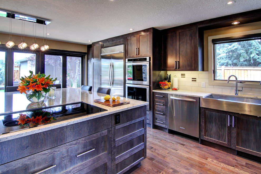 Large transitional l-shaped eat-in kitchen in Calgary with an undermount sink, shaker cabinets, dark wood cabinets, quartzite benchtops, white splashback, ceramic splashback, stainless steel appliances, medium hardwood floors, with island and brown floor.
