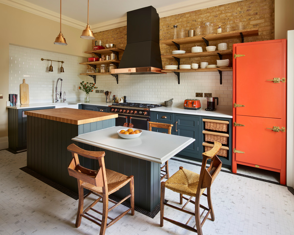This is an example of a country l-shaped kitchen in London with a farmhouse sink, recessed-panel cabinets, green cabinets, wood benchtops, white splashback, subway tile splashback, black appliances, with island, white floor and brown benchtop.