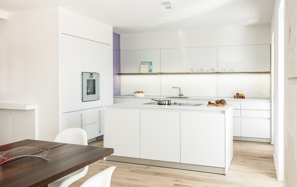 This is an example of a mid-sized contemporary single-wall eat-in kitchen in Stuttgart with a drop-in sink, flat-panel cabinets, white cabinets, white splashback, glass sheet splashback, stainless steel appliances, medium hardwood floors, with island and brown floor.