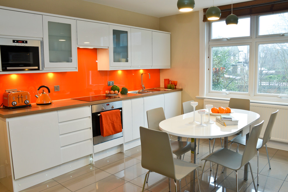 Design ideas for a mid-sized contemporary l-shaped eat-in kitchen in London with flat-panel cabinets, white cabinets, wood benchtops, orange splashback, glass sheet splashback, stainless steel appliances, porcelain floors and no island.