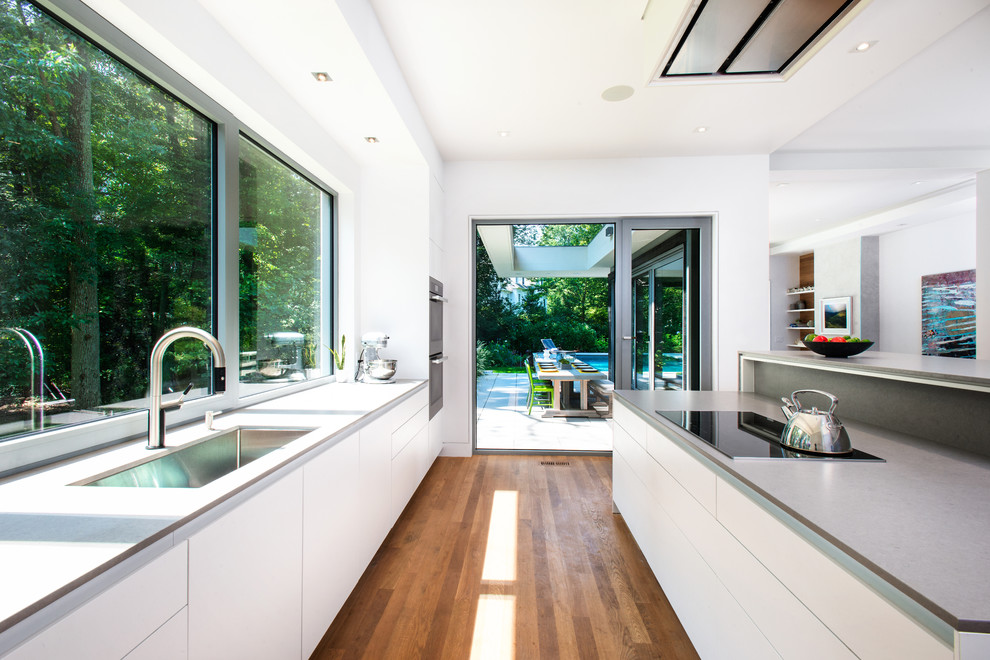 This is an example of a mid-sized contemporary single-wall open plan kitchen in New York with white cabinets, grey benchtop, a single-bowl sink, flat-panel cabinets, quartz benchtops, window splashback, light hardwood floors, with island and brown floor.