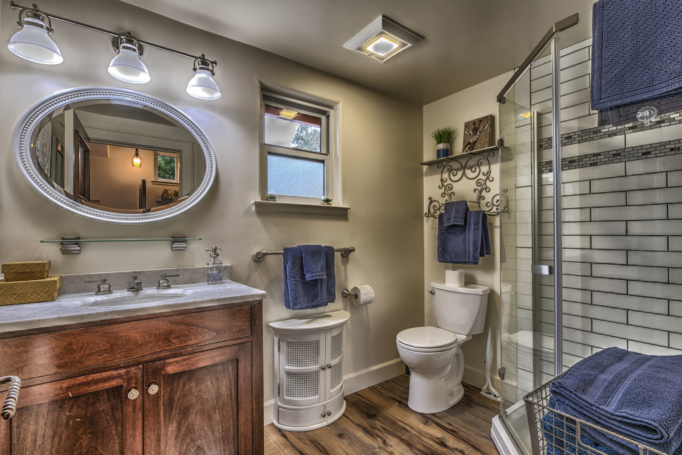 Small beach style master bathroom in San Francisco with recessed-panel cabinets, brown cabinets, a corner shower, a two-piece toilet, white tile, glass tile, beige walls, medium hardwood floors, an undermount sink, marble benchtops, beige floor, a hinged shower door and grey benchtops.