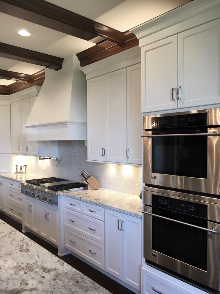 This is an example of a large transitional l-shaped eat-in kitchen in Other with a farmhouse sink, recessed-panel cabinets, white cabinets, granite benchtops, white splashback, ceramic splashback, stainless steel appliances, dark hardwood floors, with island and brown floor.