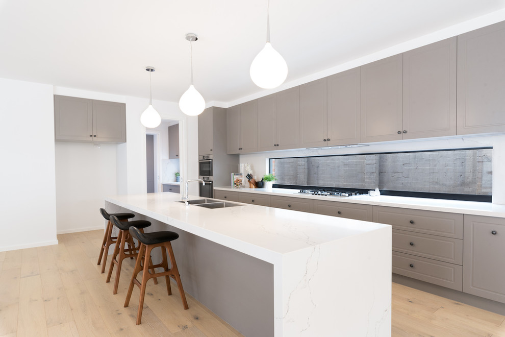 This is an example of a large contemporary galley open plan kitchen in Melbourne with a double-bowl sink, white splashback, stainless steel appliances, light hardwood floors, with island and white benchtop.