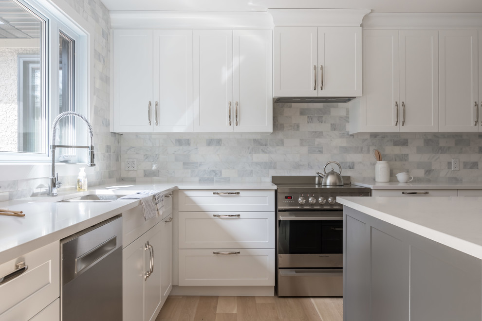 Transitional l-shaped light wood floor eat-in kitchen photo with shaker cabinets, white cabinets, solid surface countertops, white backsplash, marble backsplash, stainless steel appliances, an island and white countertops