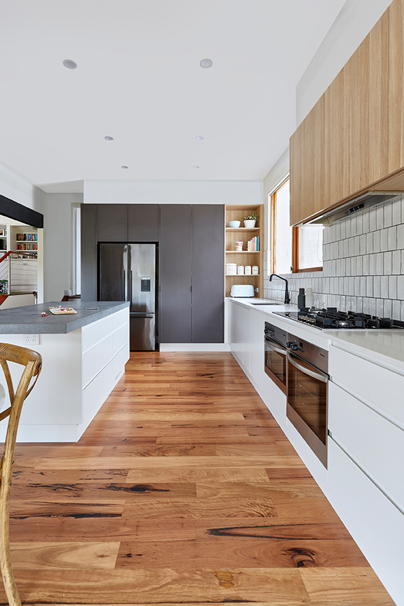 This is an example of a large contemporary galley kitchen pantry in Melbourne with a drop-in sink, raised-panel cabinets, white cabinets, solid surface benchtops, white splashback, subway tile splashback, black appliances, plywood floors, with island and beige benchtop.
