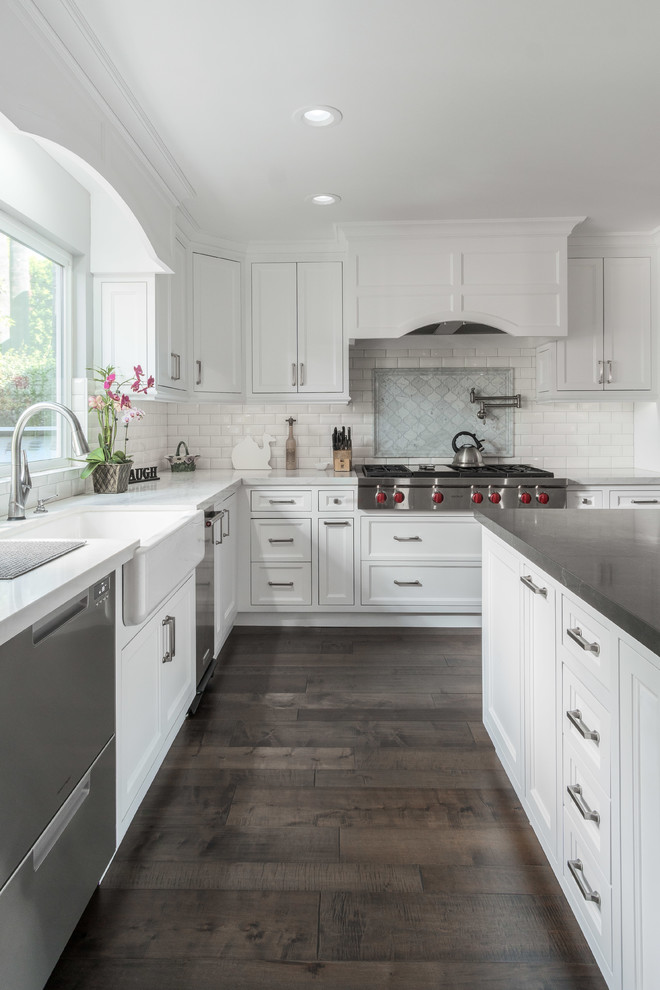 This is an example of a transitional l-shaped kitchen in Los Angeles with a farmhouse sink, recessed-panel cabinets, white cabinets, white splashback, subway tile splashback, stainless steel appliances, dark hardwood floors, with island, brown floor and white benchtop.