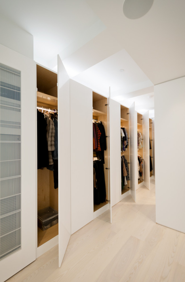 Photo of a large modern gender-neutral built-in wardrobe in New York with flat-panel cabinets, white cabinets, light hardwood floors and beige floor.