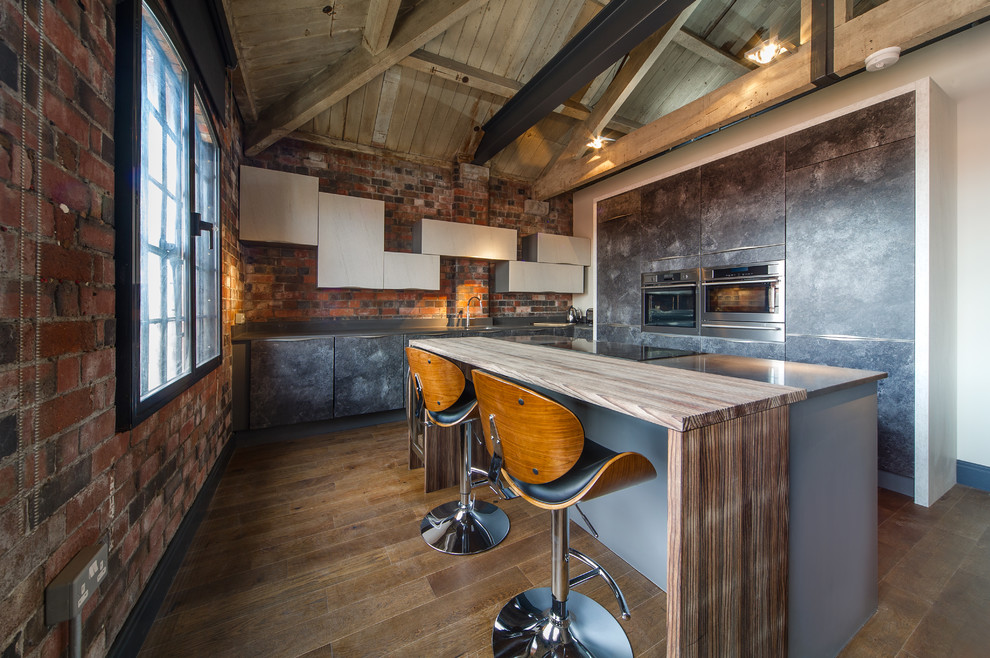 Photo of a small industrial u-shaped open plan kitchen in West Midlands with flat-panel cabinets, distressed cabinets, wood benchtops, brick splashback, stainless steel appliances, medium hardwood floors, with island and brown floor.