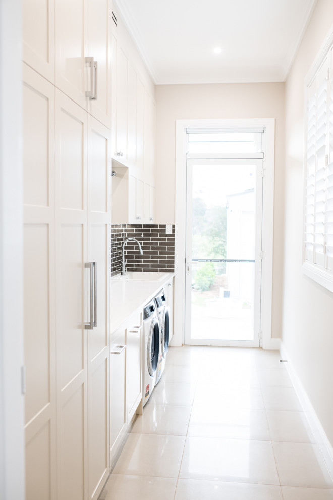 Photo of a large modern single-wall dedicated laundry room in Sydney with a drop-in sink, shaker cabinets, white cabinets, quartz benchtops, brown splashback, subway tile splashback, white walls, porcelain floors, a side-by-side washer and dryer, white floor and white benchtop.