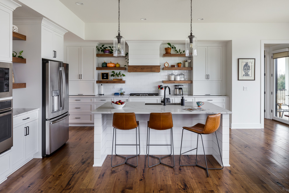 Example of a transitional medium tone wood floor and brown floor kitchen design with a farmhouse sink, shaker cabinets, white cabinets, white backsplash, subway tile backsplash, stainless steel appliances, an island and white countertops