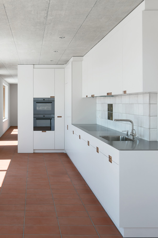 Design ideas for a mid-sized industrial l-shaped kitchen in Other with a single-bowl sink, flat-panel cabinets, white cabinets, stainless steel benchtops, white splashback, terra-cotta splashback, stainless steel appliances, terra-cotta floors, red floor and grey benchtop.