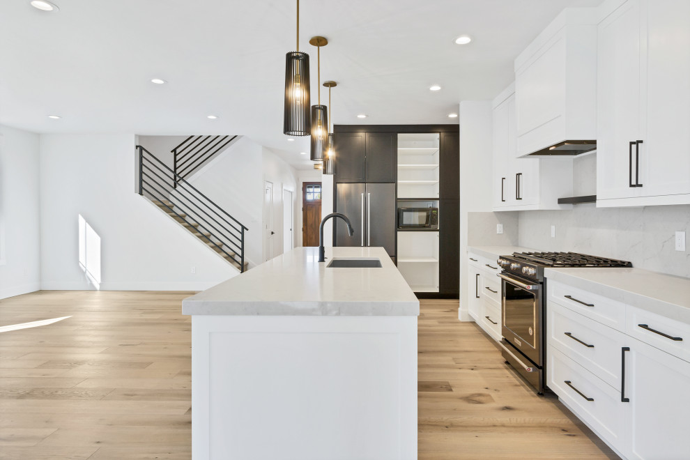 Inspiration for a mid-sized contemporary l-shaped eat-in kitchen in Other with a drop-in sink, raised-panel cabinets, white cabinets, quartz benchtops, grey splashback, subway tile splashback, black appliances, light hardwood floors, with island, beige floor and grey benchtop.