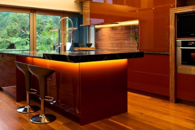 Large modern l-shaped eat-in kitchen in Auckland with multi-coloured splashback, glass sheet splashback, black appliances, with island, an undermount sink, flat-panel cabinets, red cabinets, granite benchtops and light hardwood floors.