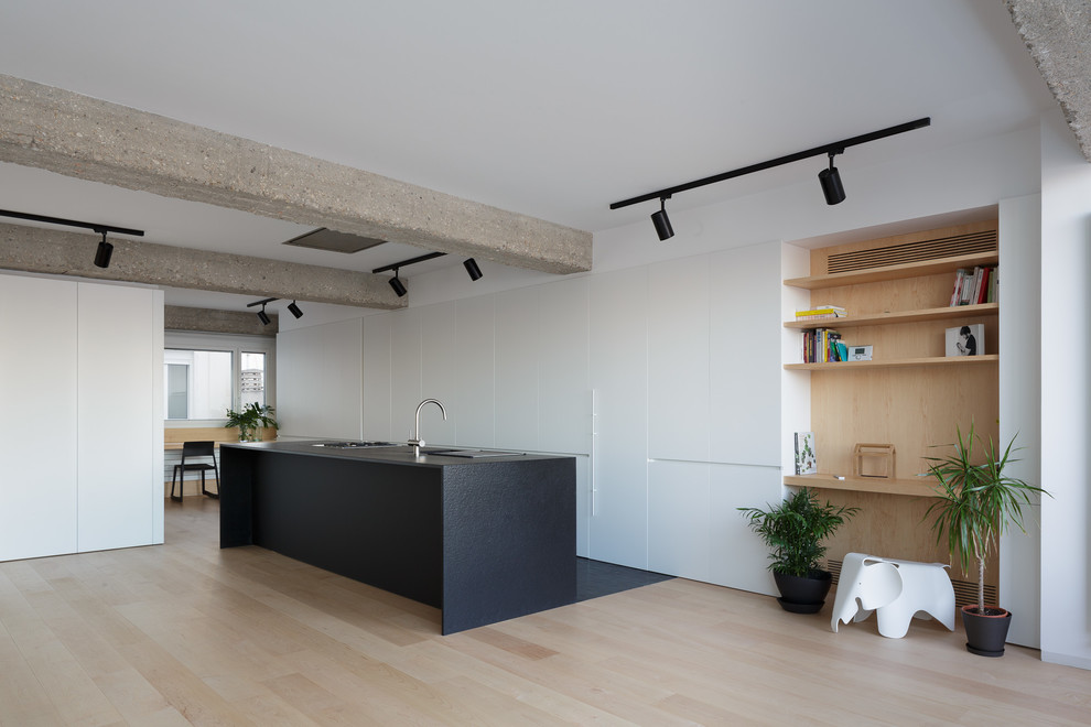 Photo of a modern single-wall open plan kitchen in Alicante-Costa Blanca with a drop-in sink, flat-panel cabinets, white cabinets, granite benchtops, black appliances, light hardwood floors, with island, black benchtop and beige floor.