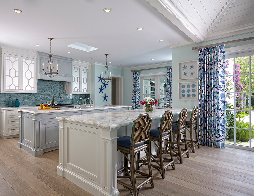 Beach style single-wall open plan kitchen in Miami with white cabinets, marble benchtops, blue splashback, mosaic tile splashback, light hardwood floors, multiple islands and recessed-panel cabinets.