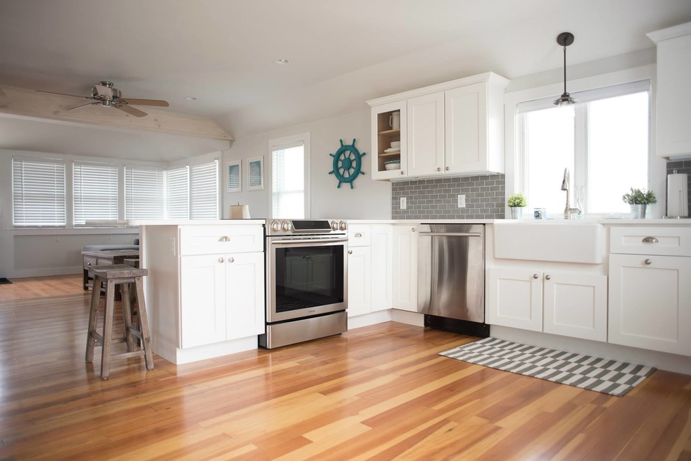 This is an example of a mid-sized beach style l-shaped eat-in kitchen in Providence with a farmhouse sink, shaker cabinets, white cabinets, quartz benchtops, grey splashback, subway tile splashback, stainless steel appliances, light hardwood floors and a peninsula.