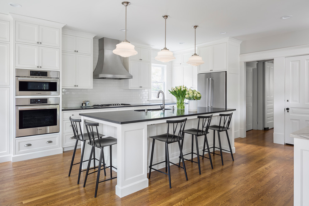 This is an example of a large transitional l-shaped kitchen in Minneapolis with an undermount sink, shaker cabinets, white cabinets, granite benchtops, white splashback, ceramic splashback, stainless steel appliances, medium hardwood floors, with island, brown floor and black benchtop.