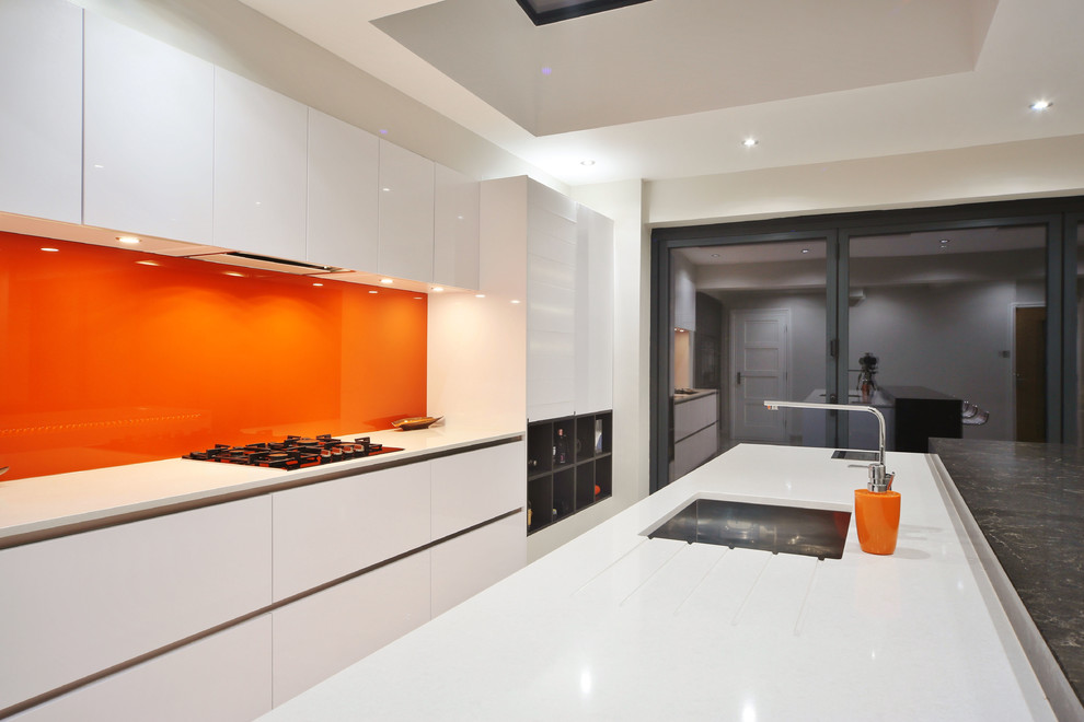 Photo of a mid-sized contemporary single-wall open plan kitchen in London with an undermount sink, flat-panel cabinets, white cabinets, quartzite benchtops, orange splashback, glass sheet splashback, black appliances, porcelain floors, with island, beige floor and white benchtop.