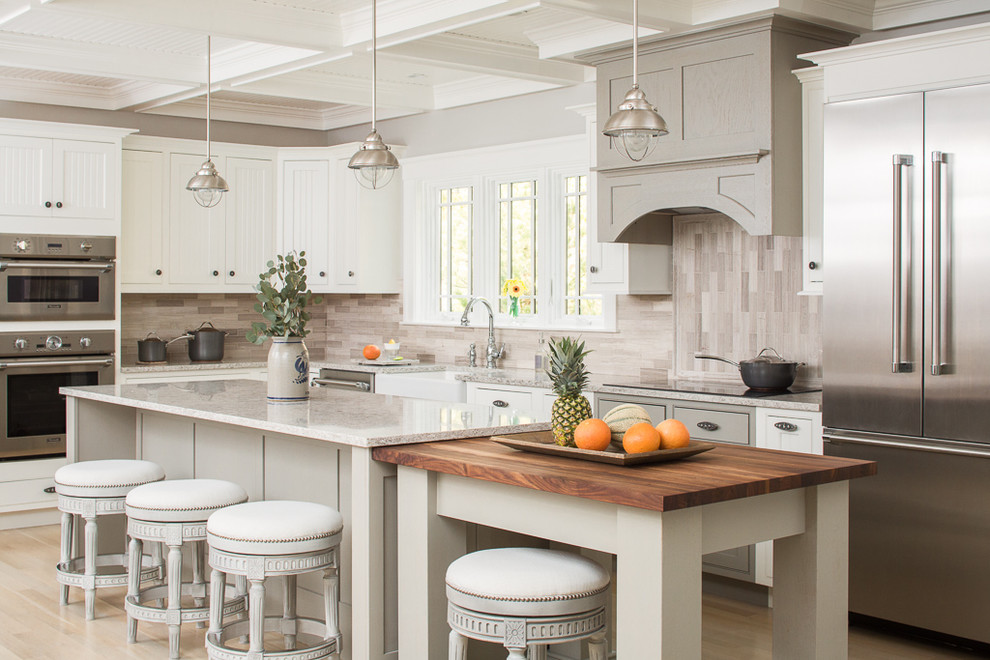 Photo of a beach style l-shaped kitchen in Portland Maine with white cabinets, a farmhouse sink, recessed-panel cabinets, wood benchtops, beige splashback, stainless steel appliances, light hardwood floors, multiple islands and beige floor.