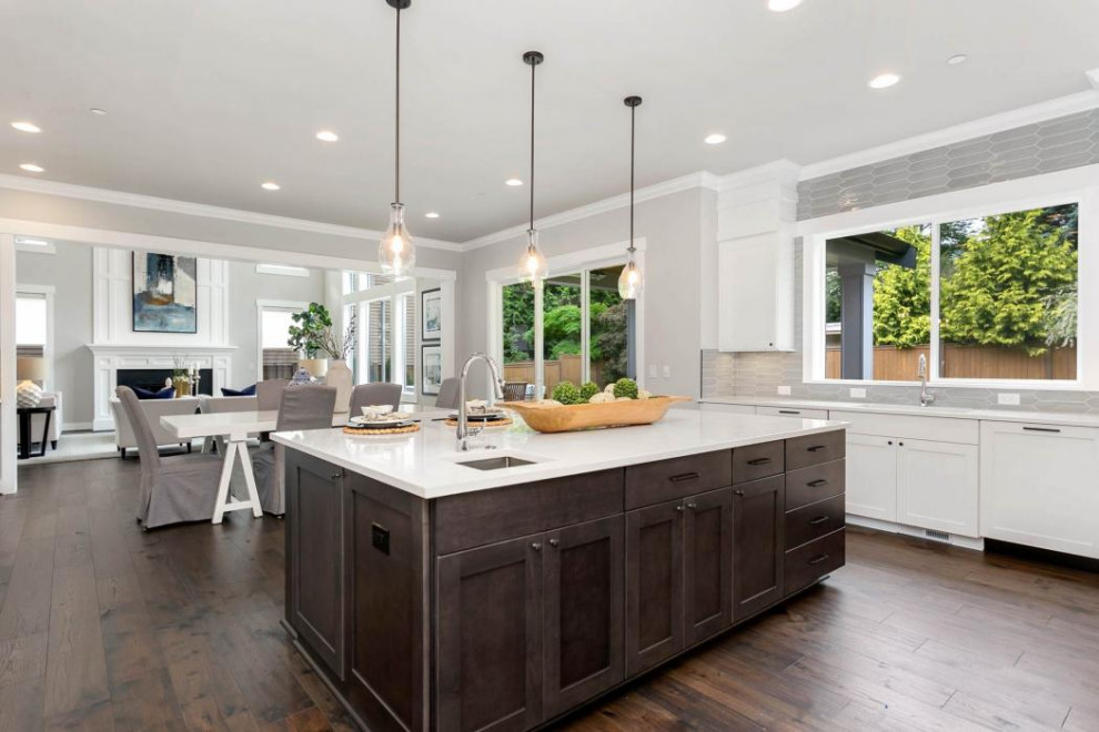 This is an example of an expansive arts and crafts u-shaped open plan kitchen in Seattle with a drop-in sink, shaker cabinets, white cabinets, quartz benchtops, grey splashback, ceramic splashback, stainless steel appliances, dark hardwood floors, with island, brown floor and white benchtop.