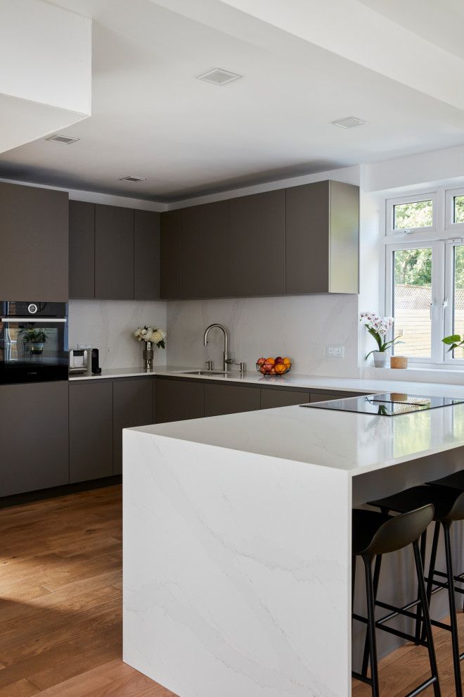 This is an example of a mid-sized modern u-shaped open plan kitchen in London with an undermount sink, flat-panel cabinets, grey cabinets, quartz benchtops, white splashback, engineered quartz splashback, stainless steel appliances, with island and white benchtop.