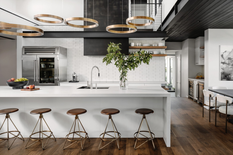 Photo of a contemporary galley kitchen/diner in Austin with a submerged sink, shaker cabinets, white cabinets, white splashback, stainless steel appliances, dark hardwood flooring, an island, brown floors and white worktops.
