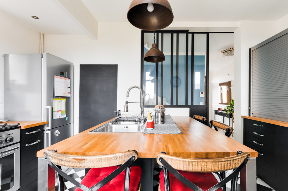 Mid-sized industrial galley separate kitchen in Paris with a double-bowl sink, shaker cabinets, black cabinets, wood benchtops, white splashback, subway tile splashback, stainless steel appliances, a peninsula, ceramic floors, black floor and brown benchtop.